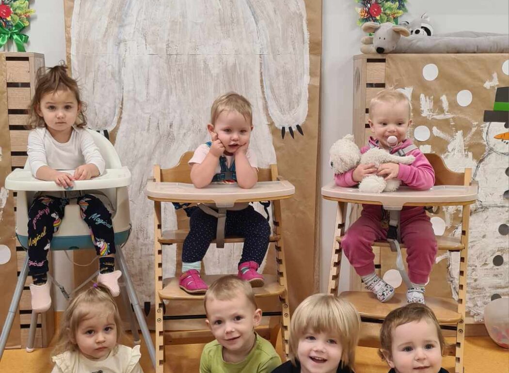 Dzieci siedzą w dwóch rzędach. W pierwszym rzędzie dzieci siedzą na podłodze, w drugim rzędzie dzieci siedzą w wysokich krzesełkach. W tle widać dużego niedźwiedzia polarnego namalowanego farbami na szarym papierze.