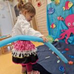 Dziewczynka stoi i trzyma w dłoniach wędkę zrobioną z podłużnego błękitnego balona. Przed nią na niebieskim materiale rozłożonym na podłodze leżą drewniane kolorowe rybki. W tle widać morskie dekoracje.