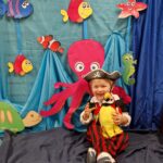 Chłopiec ubrany w strój pirata siedzi na podłodze, trzyma w dłoniach maskotkę - papugę i uśmiecha się do zdjęcia. Za chłopcem widać morskie dekoracje zawieszone na ścianie : ośmiornicę, żółwia i kolorowe rybki na niebieskim tle.