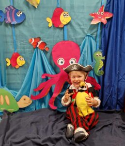 Chłopiec ubrany w strój pirata siedzi na podłodze, trzyma w dłoniach maskotkę - papugę i uśmiecha się do zdjęcia. Za chłopcem widać morskie dekoracje zawieszone na ścianie : ośmiornicę, żółwia i kolorowe rybki na niebieskim tle.