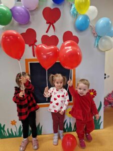 Trzy dziewczynki stoją obok siebie. Dziewczynki są ubrane na czerwono i trzymają w dłoniach czerwone balony w kształcie serc na patyczkach. W tle widać szary domek ozdobiony czerwonymi serduszkami i kolorowymi balonami.