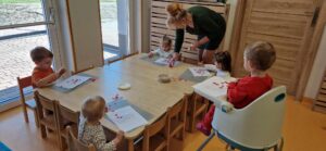 Dzieci siedzą na krzesełkach przy stole, przyklejają czerwone serduszka na białą kartkę papieru. Dzieciom pomaga opiekunka.