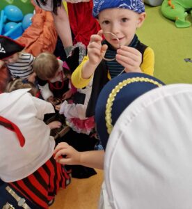 Dzieci ubrane w stroje piratów stoją wokół drewnianej skrzynki i oglądają znajdującą się w niej złotą biżuterię.