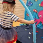 Dziewczynka ubrana w strój pirata stoi i trzyma w dłoni wędkę zrobioną z podłużnego błękitnego balona. Przed nią na niebieskim materiale rozłożonym na podłodze leżą drewniane kolorowe rybki. W tle widać morskie dekoracje.