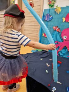 Dziewczynka ubrana w strój pirata stoi i trzyma w dłoni wędkę zrobioną z podłużnego błękitnego balona. Przed nią na niebieskim materiale rozłożonym na podłodze leżą drewniane kolorowe rybki. W tle widać morskie dekoracje.