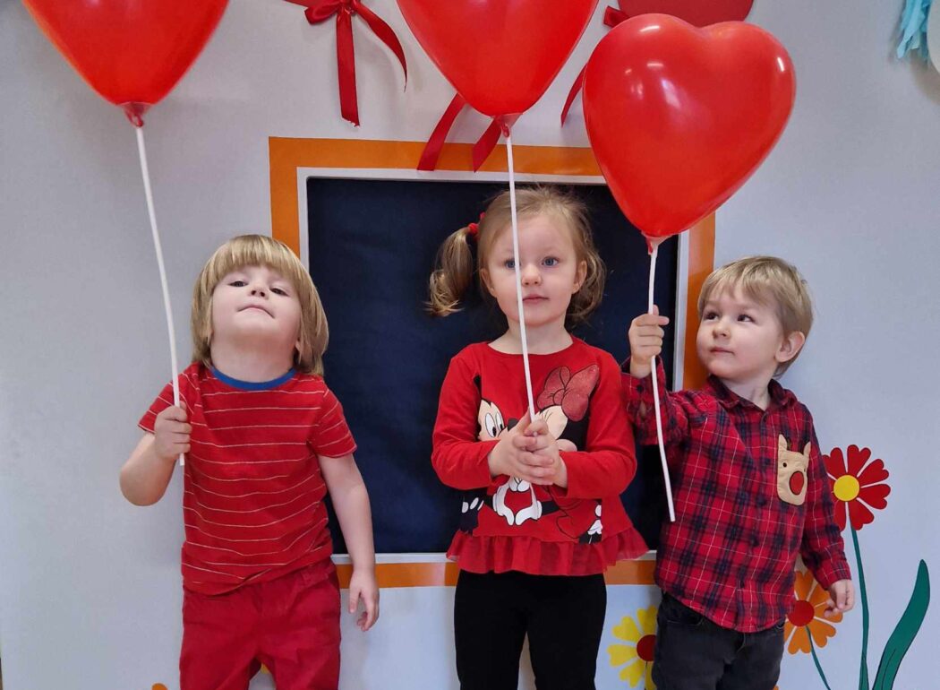 Troje dzieci stoi obok siebie. Dzieci są ubrane na czerwono i trzymają w dłoniach czerwone balony w kształcie serc na patyczkach. W tle widać szary domek ozdobiony czerwonymi serduszkami i kolorowymi balonami.