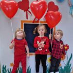 Troje dzieci stoi obok siebie. Dzieci są ubrane na czerwono i trzymają w dłoniach czerwone balony w kształcie serc na patyczkach. W tle widać szary domek ozdobiony czerwonymi serduszkami i kolorowymi balonami.