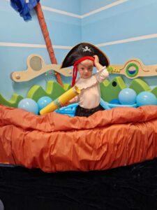 Dziewczynka ubrana w strój pirata stoi na statku zrobionym z szarego papieru. Statek jest wypełniony błękitnymi balonami. Dziewczynka trzyma w dłoniach zrobioną z papieru żółtą lunetę.