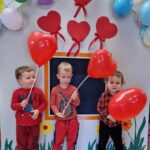 Trzech chłopców stoi obok siebie. Chłopcy są ubrani na czerwono i trzymają w dłoniach czerwone balony w kształcie serc na patyczkach. W tle widać szary domek ozdobiony czerwonymi serduszkami i kolorowymi balonami.