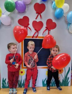 Trzech chłopców stoi obok siebie. Chłopcy są ubrani na czerwono i trzymają w dłoniach czerwone balony w kształcie serc na patyczkach. W tle widać szary domek ozdobiony czerwonymi serduszkami i kolorowymi balonami.