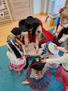 Opiekunka ubrana w strój pirata siedzi na podłodze i trzyma w dłoniach mapę. Dzieci ubrane w stroje piratów patrzą na mapę.
