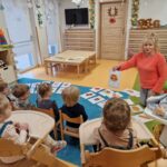 Dzieci siedzą na krzesełkach. Na dywanie leżą obrazki przedstawiające zdrową i niezdrową żywność. Opiekunka siedzi na dywanie naprzeciwko dzieci i pokazuje im jeden z obrazków.