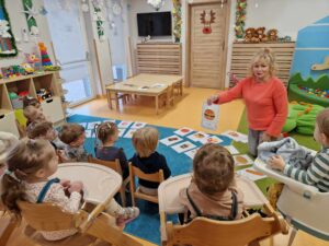 Dzieci siedzą na krzesełkach. Na dywanie leżą obrazki przedstawiające zdrową i niezdrową żywność. Opiekunka siedzi na dywanie naprzeciwko dzieci i pokazuje im jeden z obrazków.