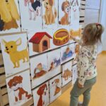 Dziewczynka stoi i patrzy na obrazki przyklejone na ścianie, przedstawiające różne rasy psów i psie akcesoria.