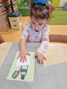 Dziewczynka siedzi przy stoliku, trzyma dłoń na kartce papieru na której namalowana jest szczoteczka i pasta do zębów.