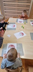 Dzieci siedzą przy stole i przyklejają smutne lub wesołe minki na twarz narysowaną na kartce papieru.
