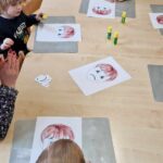 Dzieci siedzą przy stole i przyklejają smutne lub wesołe minki na twarz narysowaną na kartce papieru.