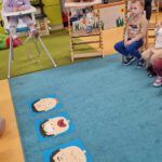 Dzieci siedzą na krzesełkach, patrzą na kolorowe obrazki rozłożone na dywanie. Obrazki przedstawiają twarze wyrażające różne emocje.