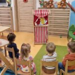 Dzieci siedzą w rzędzie na krzesełkach, oglądają przedstawienie w drewnianym teatrzyku. Teatrzyk jest ozdobiony biało-czerwonym materiałem. W tle widać salę zabaw.