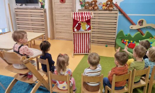 Dzieci siedzą w rzędzie na krzesełkach, oglądają przedstawienie w drewnianym teatrzyku. Teatrzyk jest ozdobiony biało-czerwonym materiałem. W tle widać salę zabaw.