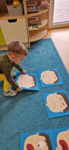 Chłopiec układa na turkusowym dywanie kolorowe obrazki. Na obrazkach namalowane są twarze, wyrażające różne emocje.