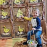 Chłopiec siedzi na krzesełku, w dłoni trzyma koszyk z jajkami. W tle widać kurnik zrobiony z szarego papieru, z żółtymi kurkami na grzędach.