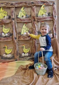 Chłopiec siedzi na krzesełku, w dłoni trzyma koszyk z jajkami. W tle widać kurnik zrobiony z szarego papieru, z żółtymi kurkami na grzędach.