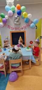 Dzieci ubrane w kolorowe, urodzinowe czapeczki siedzą na krzesełkach przy stole i jedzą babeczki. W tle widać szary domek ozdobiony urodzinowymi dekoracjami: różowym napisem, srebrnym balonem w kształcie cyfry dwa i kolorowymi balonikami.