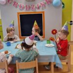 Dzieci ubrane w kolorowe, urodzinowe czapeczki siedzą na krzesełkach przy stole i jedzą babeczki. W tle widać szary domek ozdobiony urodzinowymi dekoracjami: różowym napisem, srebrnym balonem w kształcie cyfry dwa i kolorowymi balonikami.