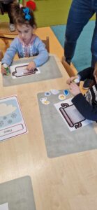 Dzieci siedzą przy stole i przyklejają małe kolorowe grafiki na garnek narysowany na kartce papieru.