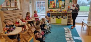 Dzieci siedzą na krzesełkach, patrzą na opiekunkę która stoi naprzeciwko i pokazuje im kolorową grafikę. Na turkusowym dywanie leżą kolorowe grafiki.