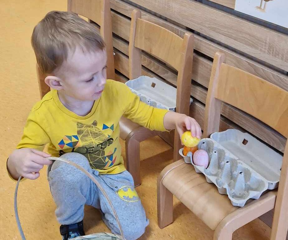 Chłopiec siedzi na podłodze, obok niego stoją trzy krzesełka ustawione pod ścianą. Chłopiec trzyma w rączce koszyk i wkłada kolorowe jajko do wytłaczanki.