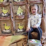 Dziewczynka siedzi na krzesełku, w rączkach trzyma koszyk z jajkami. W tle widać kurnik zrobiony z szarego papieru, z żółtymi kurami na grzędach.