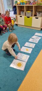 Na turkusowym dywanie leżą grafiki przedstawiające różne zjawiska atmosferyczne. Chłopiec ogląda jedną z grafik. W tle widać dzieci siedzące na krzesełkach.