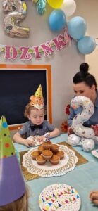 Chłopiec ubrany w kolorową, urodzinową czapkę siedzi razem z opiekunką przy stole. Na stole leży duży talerz z babeczkami i balon w kształcie cyfry dwa. W tle widać urodzinowe dekoracje.