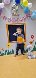 Chłopiec stoi i trzyma w dłoni żółty balon. Za chłopcem widać szary domek ozdobiony kwiatkami i urodzinowymi dekoracjami: różowym napisem, srebrnym balonem w kształcie cyfry dwa i kolorowymi balonami.