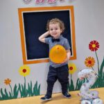 Chłopiec stoi i trzyma w dłoni żółty balon. Za chłopcem widać szary domek ozdobiony kwiatkami i urodzinowymi dekoracjami: różowym napisem, srebrnym balonem w kształcie cyfry dwa i kolorowymi balonami.