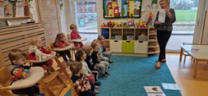 Dzieci siedzą na krzesełkach, patrzą na opiekunkę która stoi naprzeciwko i pokazuje im kolorową grafikę.