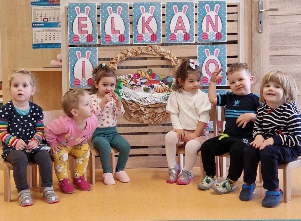 Dzieci siedzą na krzesełkach ustawionych w rzędzie. Za nimi widać przyklejony na ścianie różowo-niebieski napis "Wielkanoc" i duży wielkanocny koszyk wykonany z papieru.