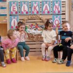 Dzieci siedzą na krzesełkach ustawionych w rzędzie. Za nimi widać przyklejony na ścianie różowo-niebieski napis "Wielkanoc" i duży wielkanocny koszyk wykonany z papieru.