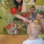 Opiekunka ubrana w kolorową, kwiecistą suknię i kapelusz ozdobiony kwiatami rozdaje tulipany dzieciom.
