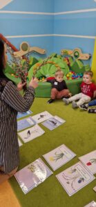 Dzieci siedzą w rzędzie na zielonym dywanie. Opiekunka trzyma w dłoniach bazie i pokazuje je dzieciom. Na dywanie przed dziećmi leżą kolorowe grafiki przedstawiające zwiastuny wiosny.