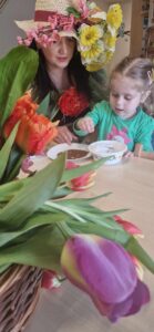 Dziewczynka wsypuje ziarenka rzeżuchy do plastikowej miseczki, pomaga jej opiekunka, która ma na głowie kapelusz ozdobiony kolorowymi kwiatami.