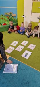 Dzieci siedzą w rzędzie na zielonym dywanie. Naprzeciwko nich siedzi opiekunka która pokazuje im rozłożone na dywanie kolorowe grafiki, przedstawiające zwiastuny wiosny.