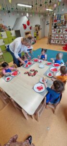 Dzieci ubrane w granatowe fartuszki siedzą przy stole i malują czerwoną farbą papierowe talerzyki. Dzieciom pomaga opiekunka.