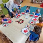 Dzieci ubrane w granatowe fartuszki siedzą przy stole i malują czerwoną farbą papierowe talerzyki. Dzieciom pomaga opiekunka.