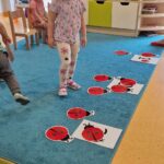 Dziewczynka stoi i patrzy na rozłożone na dywanie czerwone biedronki wykonane z papieru.