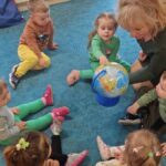 Dzieci siedzą w kole na turkusowym dywanie i patrzą na duży globus który pokazuje im opiekunka.