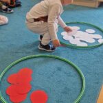 Chłopiec wkłada białe papierowe kółka do zielonej obręczy położonej na dywanie. Obok leży druga obręcz wypełniona czerwonymi kółkami.