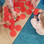 Dzieci siedzą na dywanie i wyklejają czerwonymi kółkami kontur Polski narysowany na szarym papierze. Dzieciom pomaga opiekunka.